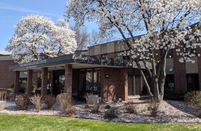 Exterior view of a healthcare facility building with brick walls and large windows. The entrance has a covered walkway supported by brick columns. There are blooming white flowering trees and landscaped bushes around the entrance area under a clear blue sky.