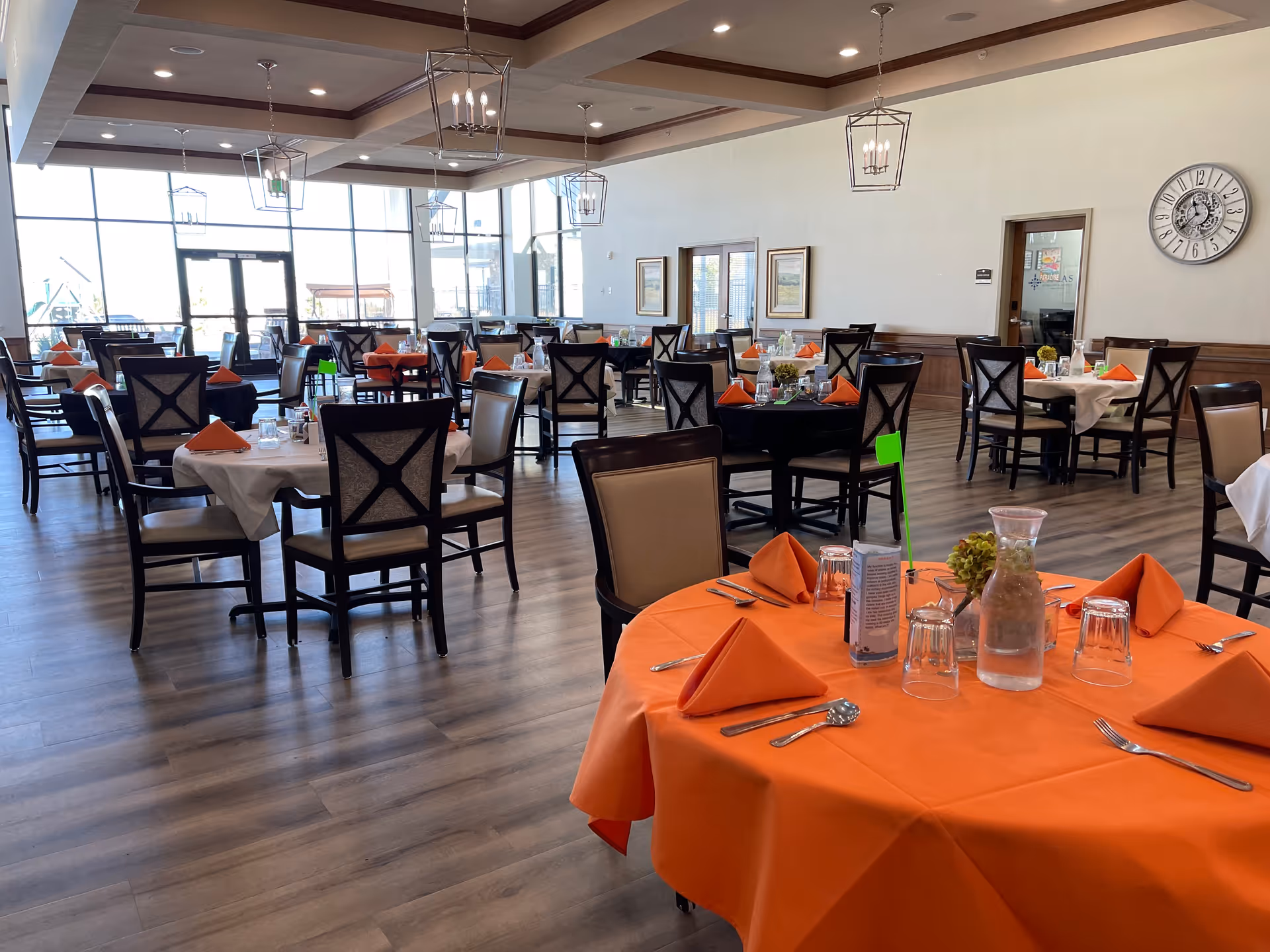 A spacious dining room with multiple round tables covered with white and orange tablecloths, each set with orange folded napkins, glasses, and silverware. The room has large windows allowing natural light, wooden flooring, and modern hanging light fixtures. There are framed pictures on the walls and a large clock.