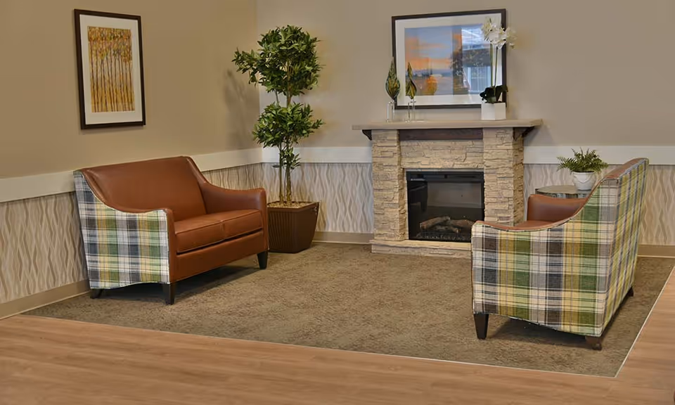 A cozy sitting area with two plaid armchairs featuring brown leather cushions, positioned around a stone fireplace. There is a potted plant in the corner and framed artwork on the walls, with a small table holding a plant beside one of the chairs.