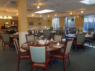 A spacious dining room with multiple round tables set with cups, plates, and utensils. Each table is surrounded by wooden chairs with green upholstered backs and seats. The room has large windows allowing natural light to enter, and ceiling lights provide additional illumination. The walls are decorated with framed pictures and the floor is carpeted.