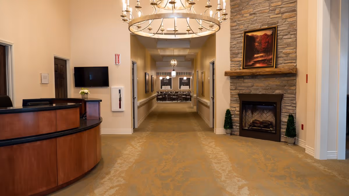 Interior view of a senior living facility hallway with a curved wooden reception desk on the left, a stone fireplace with a framed picture above it on the right, and a dining area visible at the end of the hallway. The hallway is carpeted and well-lit with chandeliers and pendant lights.