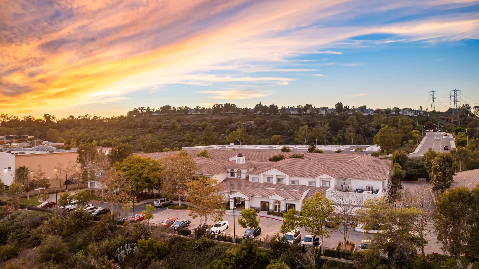 Aerial view of a senior living facility with red-tiled roofs, a parking lot and surrounding trees on a hillside at sunset.