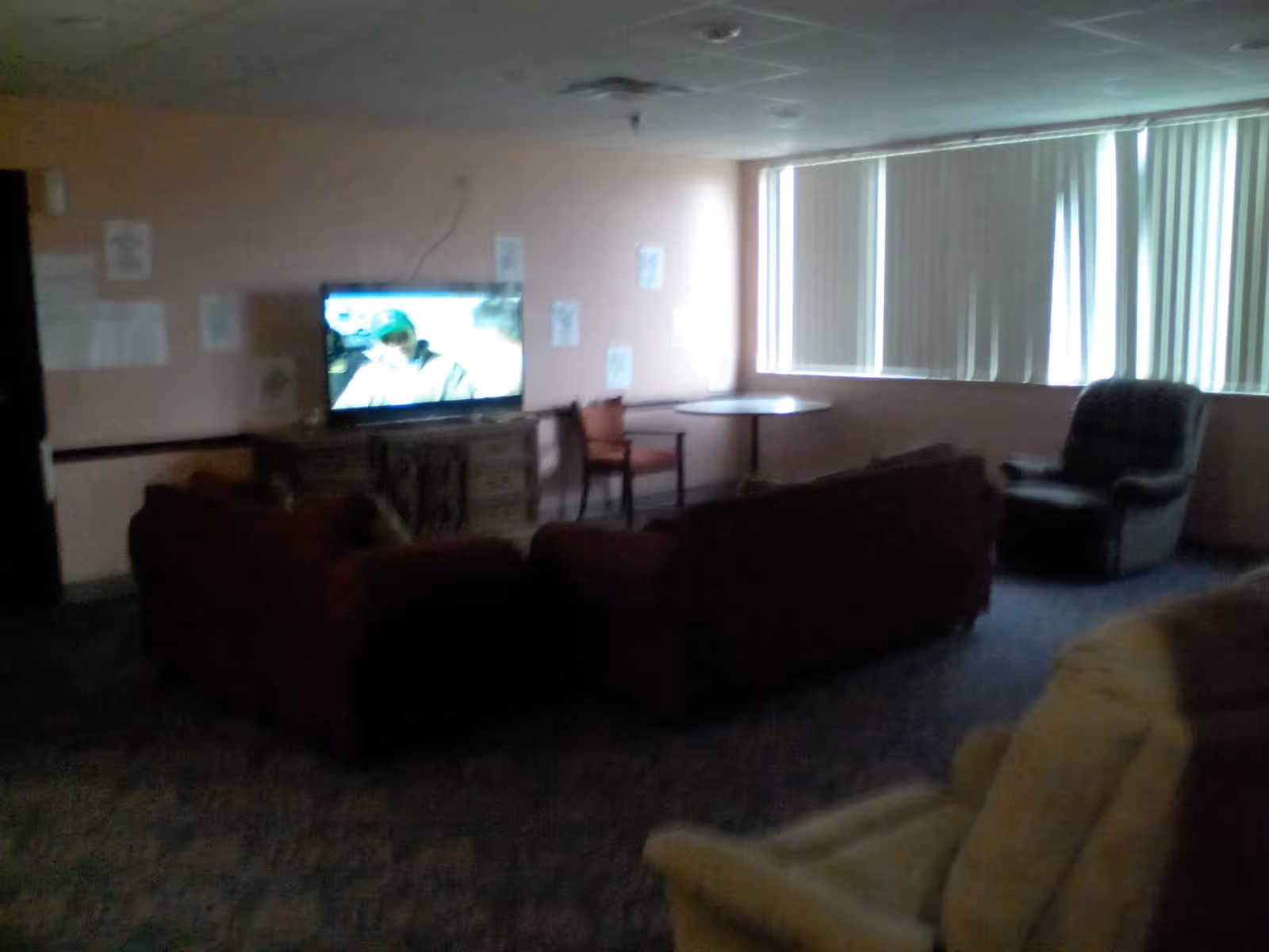 A dimly lit living room with a television on a wooden stand showing a person on screen. The room has several chairs and couches arranged facing the TV. There is a round table with two chairs near a window with vertical blinds partially closed.