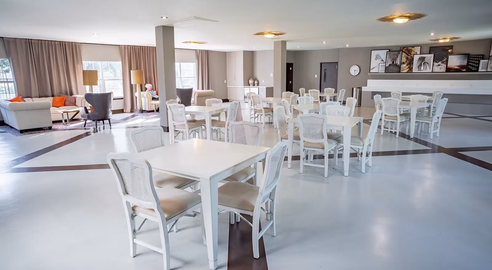 A spacious and bright common area with multiple white tables and chairs arranged neatly on a polished floor. In the background, there is a seating area with sofas, armchairs, and floor lamps near large windows with curtains. The walls have framed artwork and a clock, creating a welcoming atmosphere.