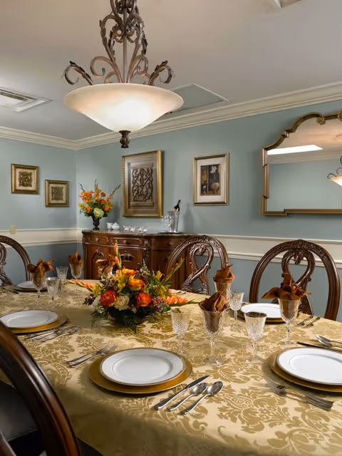 Formal dining room with an ornate chandelier, a set table with a floral centerpiece, and a wooden sideboard against a blue-gray wall.