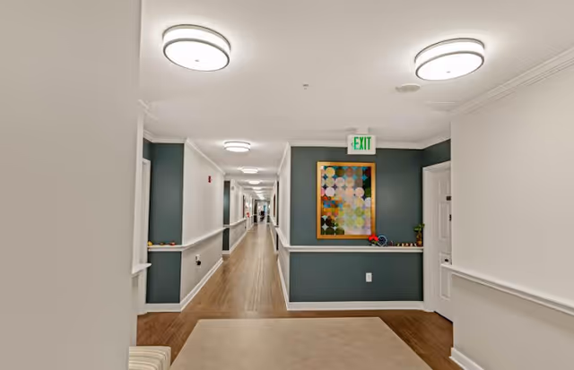 A long, well-lit hallway in a senior living facility with wooden flooring and white walls accented by teal panels. The hallway features ceiling lights, an exit sign, a colorful abstract painting on the wall, and a small ledge with decorative items.