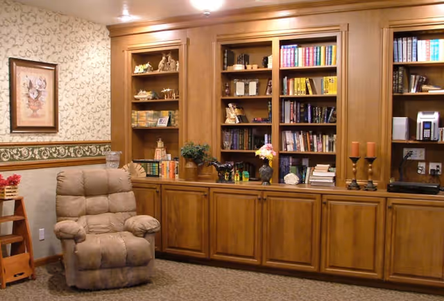 A cozy living room area with a beige recliner chair, wooden built-in bookshelves filled with books and decorative items, floral wallpaper with a framed flower painting, and a carpeted floor.