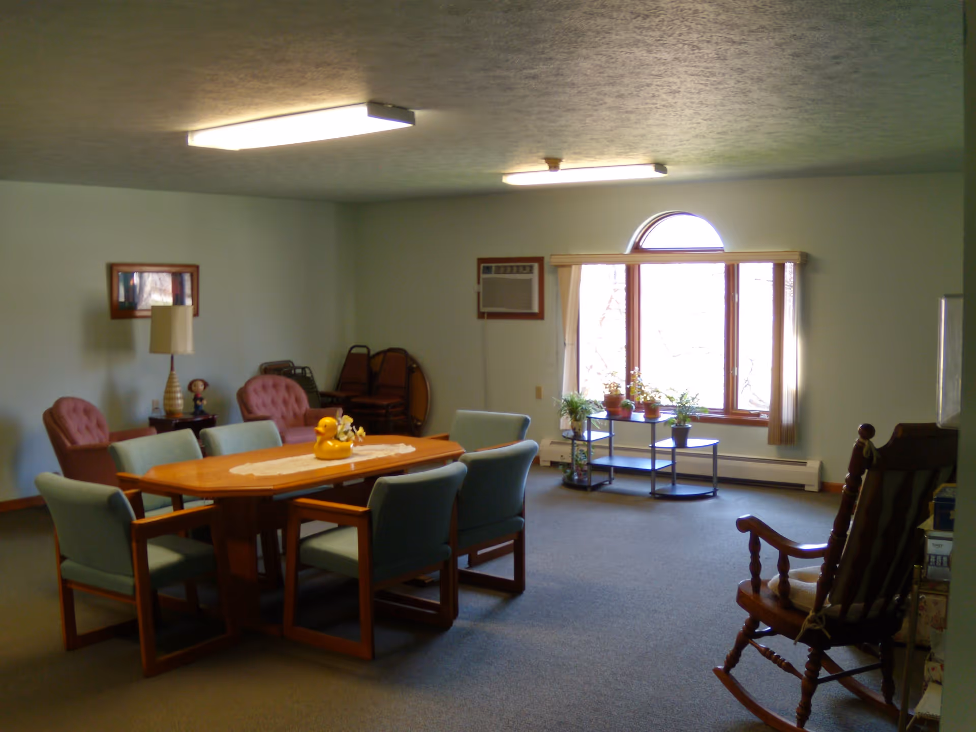 A spacious room with a wooden table surrounded by six cushioned chairs. In the background, there are two pink armchairs, a side table with a lamp and a small doll, a window with vertical blinds, and a small shelf with potted plants. A wooden rocking chair is visible in the foreground on the right side. The room has carpeted flooring and fluorescent ceiling lights.