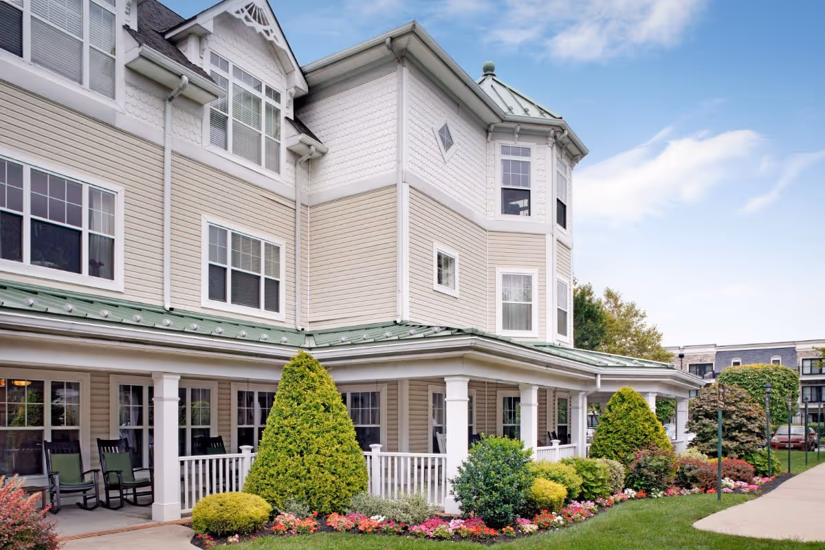 Exterior front of a multi-story senior living building with a covered porch, rocking chairs and well-kept landscaping.