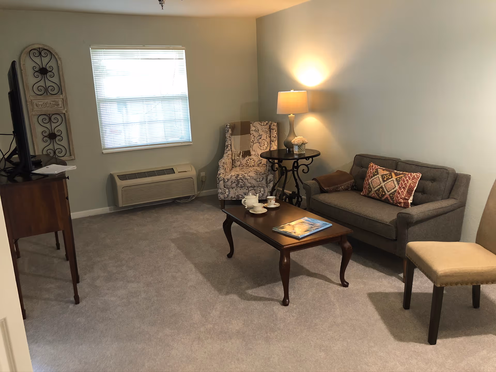 A cozy living room with a gray sofa adorned with a patterned pillow and a brown throw blanket, a floral armchair with a beige and brown throw, a wooden coffee table with a tea set and a book, a round side table with a lamp and a small flower arrangement, a beige upholstered chair, a window with blinds, and a TV on a wooden stand.