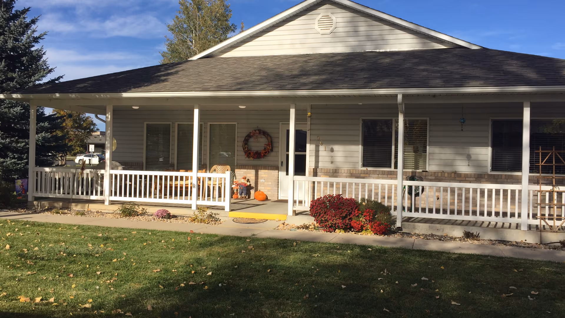 Front view of a single-story building with a covered porch, white railing, fall decorations, and a grassy lawn.