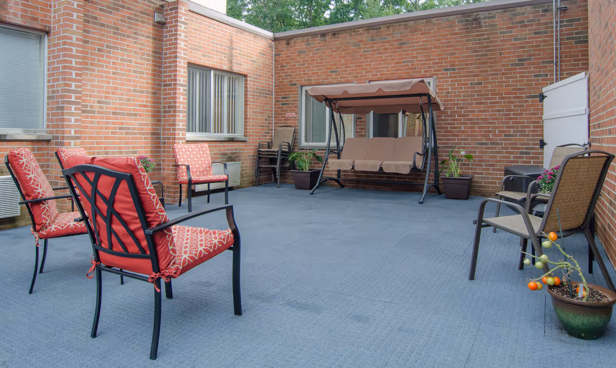 Outdoor patio area with brick walls, featuring several chairs with red cushions, a brown cushioned swing, potted plants, and a small tomato plant in a pot on the floor.