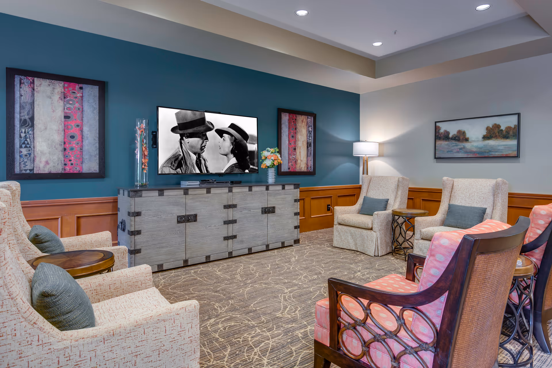 A cozy living room area with a large flat-screen TV mounted on a teal accent wall. The TV is displaying a black and white movie scene featuring a man and a woman wearing hats. The room has several upholstered armchairs with cushions arranged around small round wooden tables. There are two framed abstract artworks on the teal wall and a landscape painting on the adjacent light gray wall. A floor lamp provides soft lighting in the corner, and the floor is carpeted with a patterned design.