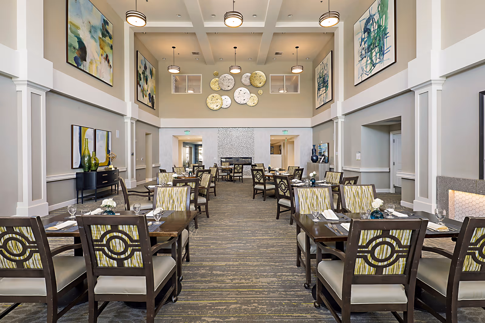High-ceiling communal dining room with neatly set tables, patterned chairs, and large wall art.