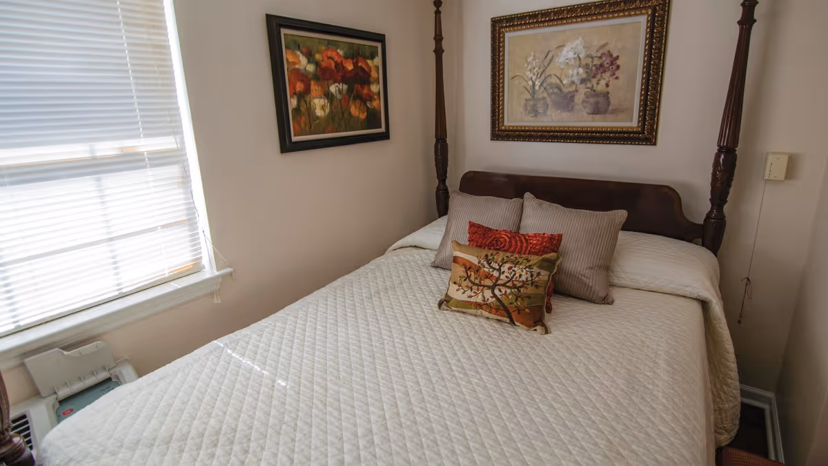 A cozy bedroom with a wooden four-poster bed covered with a white quilted bedspread and three decorative pillows. Two framed floral paintings hang on the walls above the bed. A window with closed blinds is on the left side, allowing natural light to softly illuminate the room.