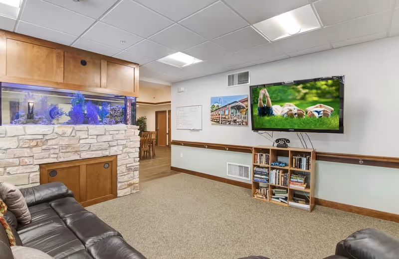 A cozy senior living common area featuring a large leather couch, a built-in aquarium with blue lighting, a wall-mounted flat-screen TV displaying an image of people lying on grass, a small bookshelf filled with books, and a whiteboard on the wall. The room has carpeted flooring and a ceiling with recessed lighting.