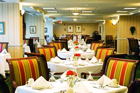 A well-lit dining room with multiple tables covered in white tablecloths, each set with folded napkins, glassware, and small floral centerpieces. The chairs have striped upholstery in shades of red, green, and yellow. The room has patterned wallpaper, framed pictures on the walls, and ceiling lights providing warm illumination.