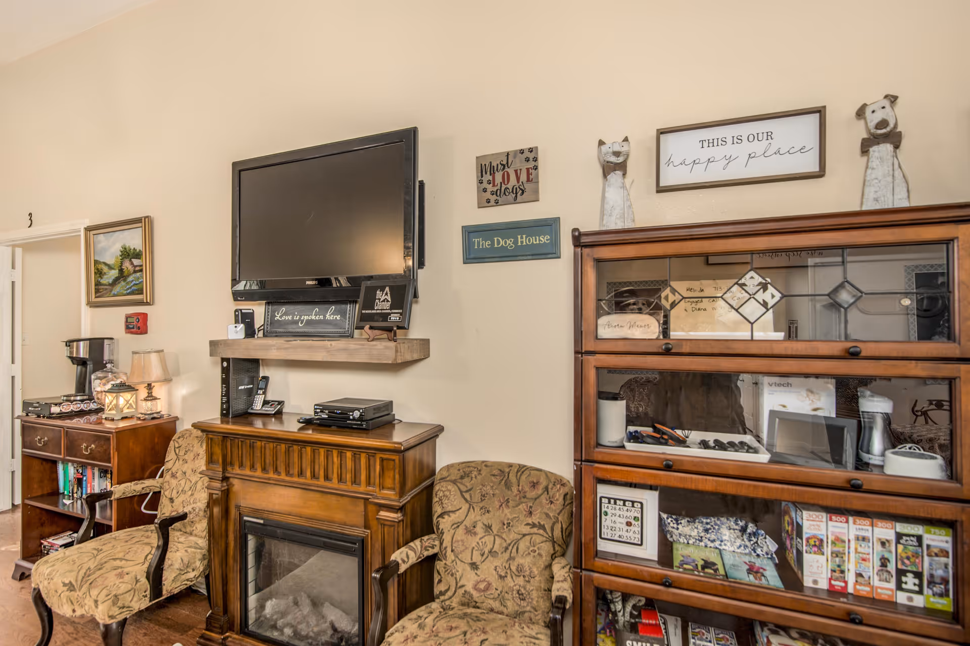 A cozy living room area with two patterned armchairs, a wooden electric fireplace with a flat-screen TV mounted above it, and a wooden cabinet filled with various items including books and games. The walls are decorated with signs that say 'Must Love Dogs,' 'The Dog House,' and 'This is Our Happy Place.' There is also a small wooden table with a lamp and coffee maker in the background.