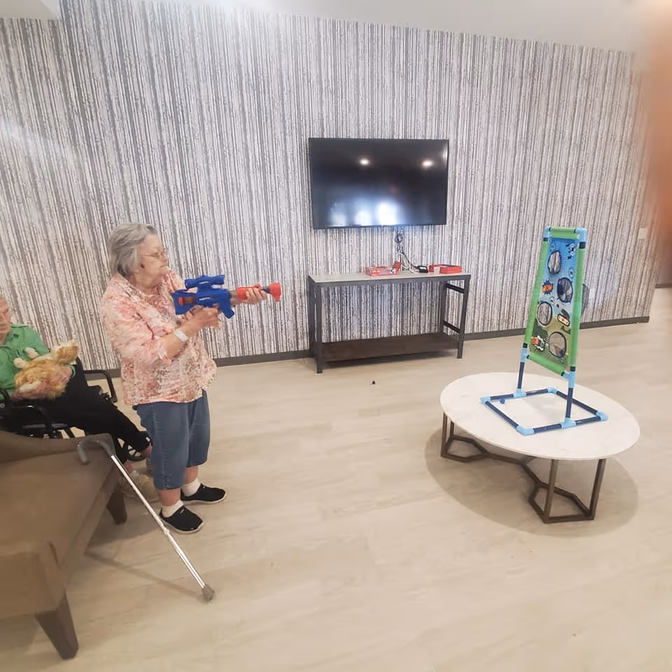 An elderly woman standing and aiming a toy gun at a target game set up on a round table in a room with light wood flooring and patterned wallpaper. Another elderly person is seated on a chair nearby holding a stuffed animal. A flat-screen TV is mounted on the wall above a small table with some items on it.