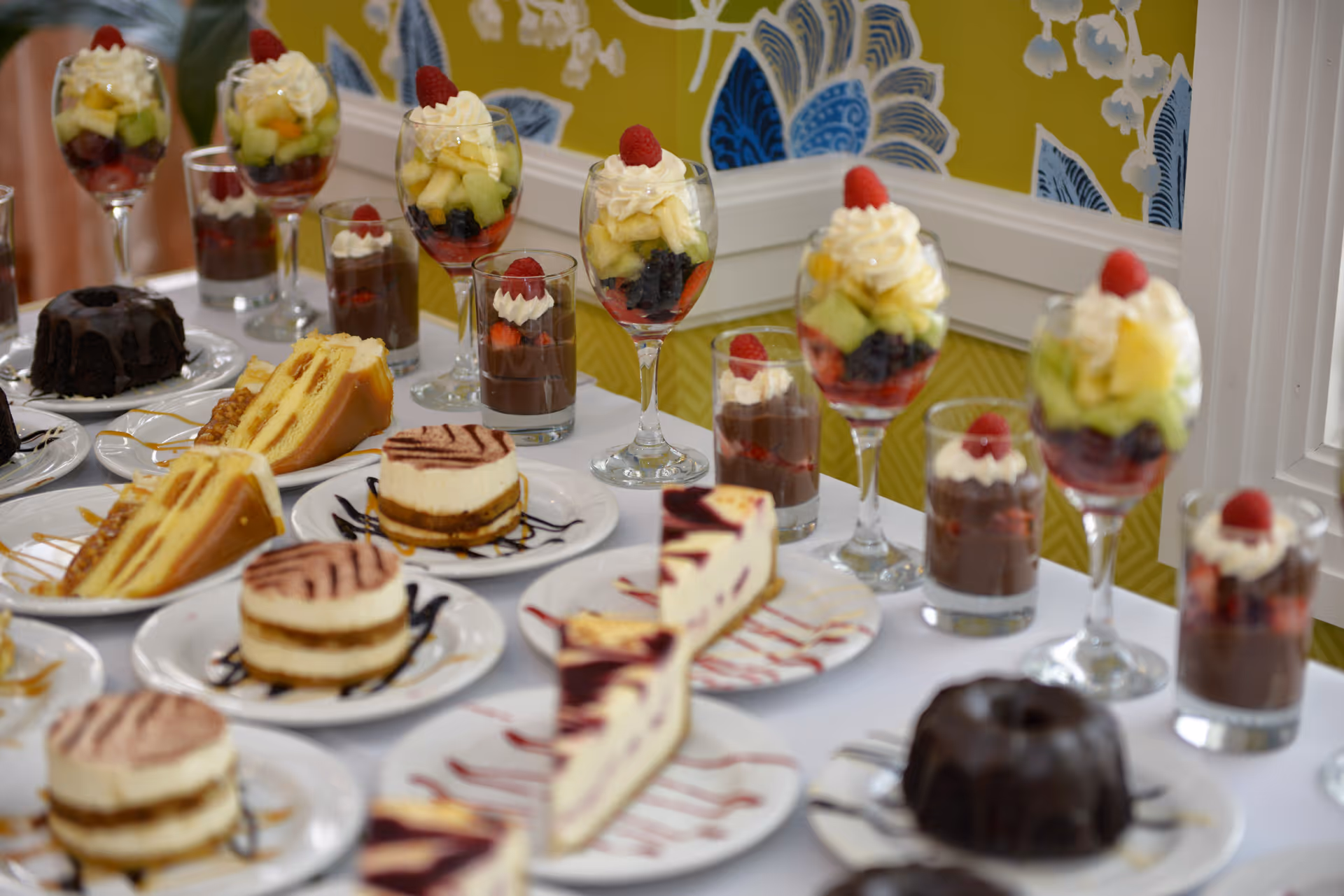 A table set with an assortment of desserts including layered cakes, chocolate mousse topped with whipped cream and raspberries, fruit parfaits in wine glasses with whipped cream and raspberries, and chocolate bundt cakes. The background features a yellow wall with blue and white floral patterns.