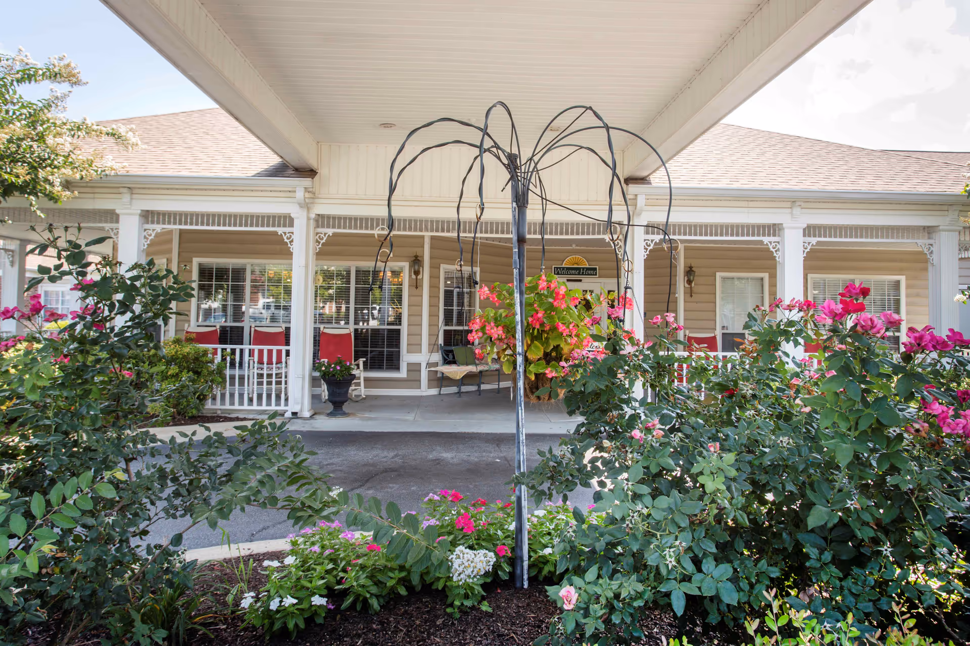 Covered entrance porch of a residential building with potted plants, red chairs, and flowering shrubs in the foreground.
