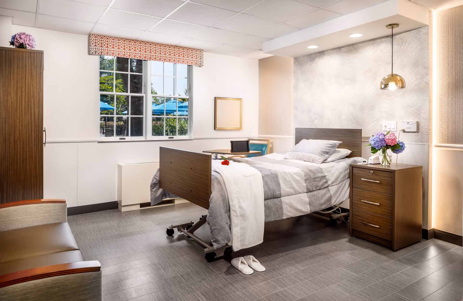 A well-lit, modern nursing facility bedroom with a single hospital bed covered in a gray and white checkered blanket. A red rose and a white towel are placed on the bed. Next to the bed is a wooden nightstand with a vase of colorful flowers and a telephone. A gold pendant light hangs above the nightstand. There is a window with a patterned valance showing an outdoor view with blue umbrellas. A small table with a chair and a tablet is positioned near the window. A cushioned armchair is partially visible on the left side of the room.