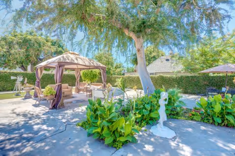 Outdoor garden area with a large tree providing shade, a gazebo with curtains and seating underneath, surrounded by green plants and bushes. There is a small white statue in the foreground and a patio umbrella with chairs in the background.