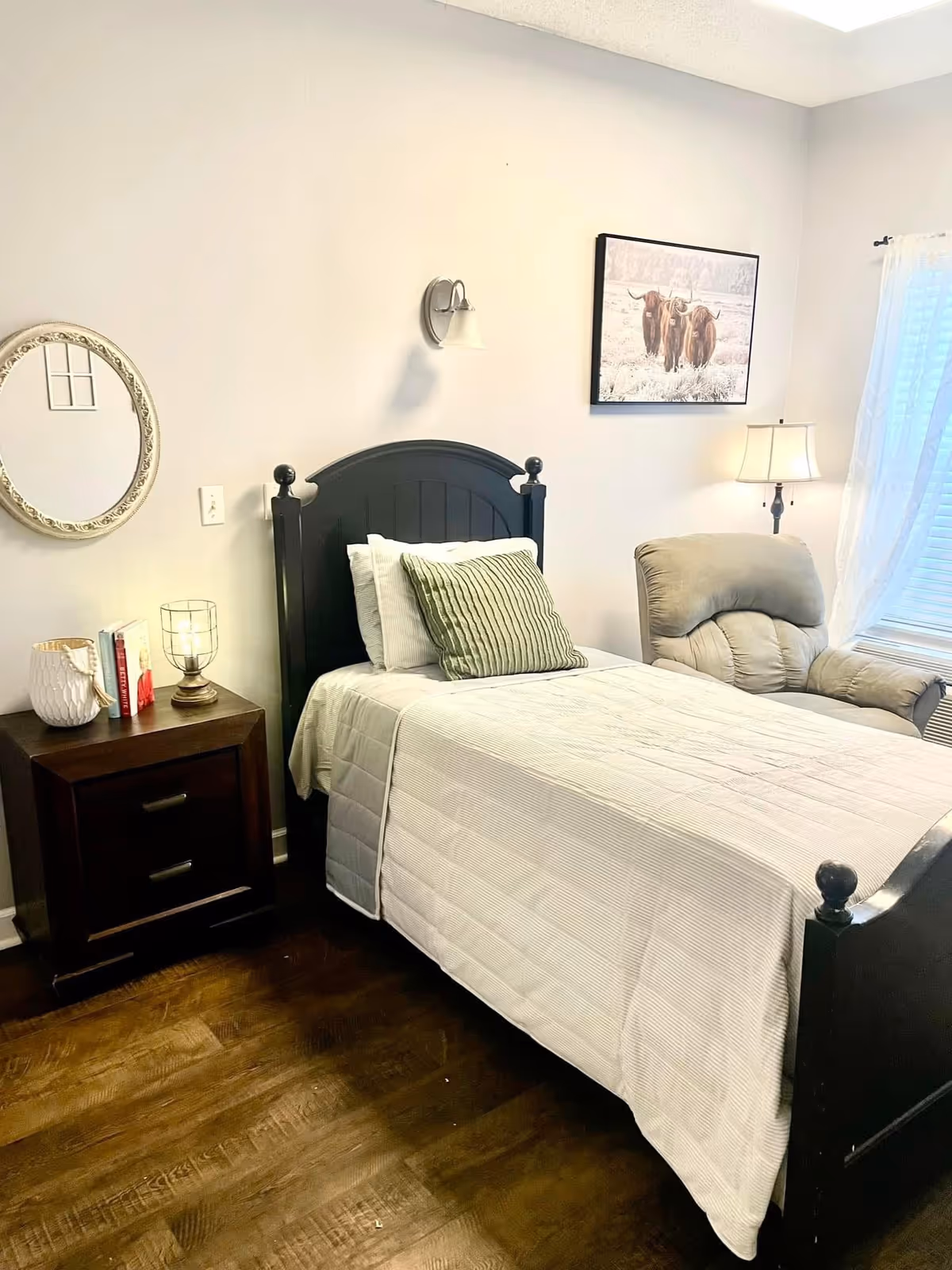A cozy bedroom with a single black wooden bed featuring white bedding and a green pillow. Next to the bed is a dark wooden nightstand with two drawers, holding a white vase, some books, and a decorative lamp. On the wall above the bed is a wall-mounted light fixture and a framed picture of two highland cows. A comfortable beige recliner chair is positioned near a window with white sheer curtains, allowing natural light to enter the room. The floor is dark wood, and the walls are painted light gray.