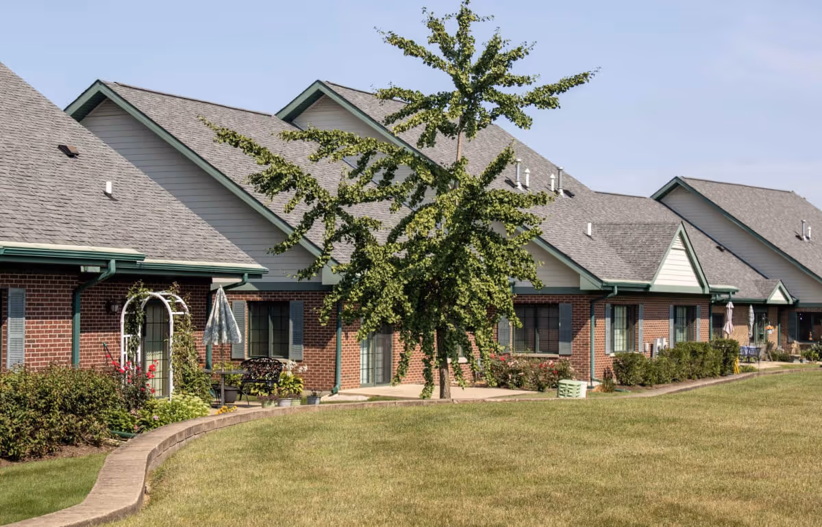 Front view of a row of single-story brick senior living units with green trim, patios, lawn and a tree.