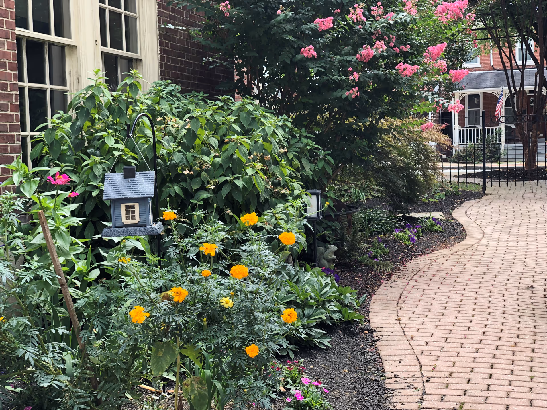 A garden area beside a brick building with green leafy plants and blooming flowers in yellow, pink, and purple. A small decorative birdhouse hangs on a metal hook among the plants. A curved brick pathway leads to a black metal gate with a view of a porch and American flags in the background.