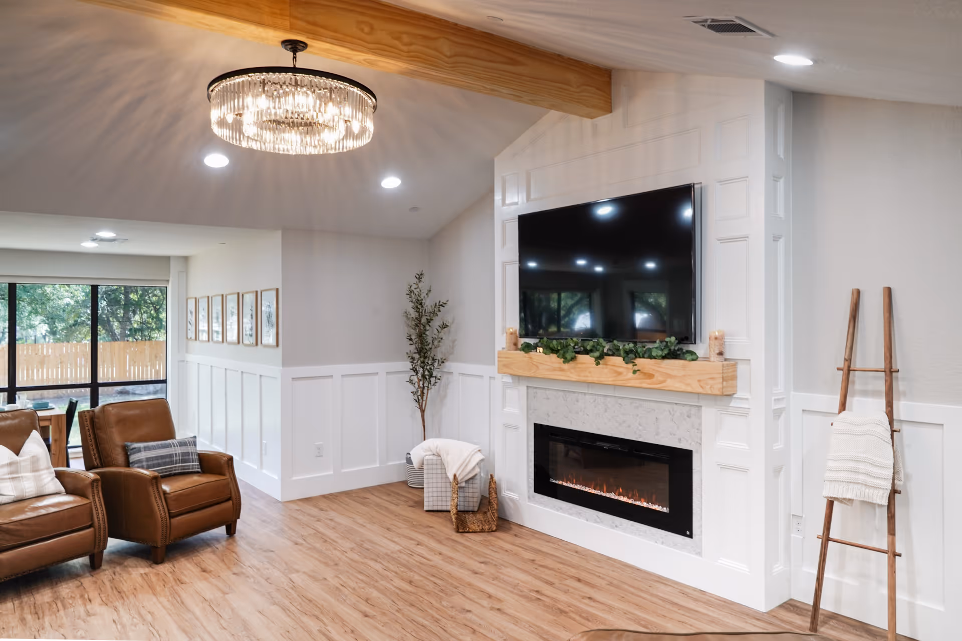 A cozy living room with two brown leather armchairs with pillows, a modern electric fireplace with a wooden mantel decorated with greenery, and a large flat-screen TV mounted above it. The room has light-colored walls with white wainscoting, a wooden floor, a decorative ceiling light fixture, and a tall plant in the corner. A wooden ladder with a white blanket is leaning against the wall on the right side.
