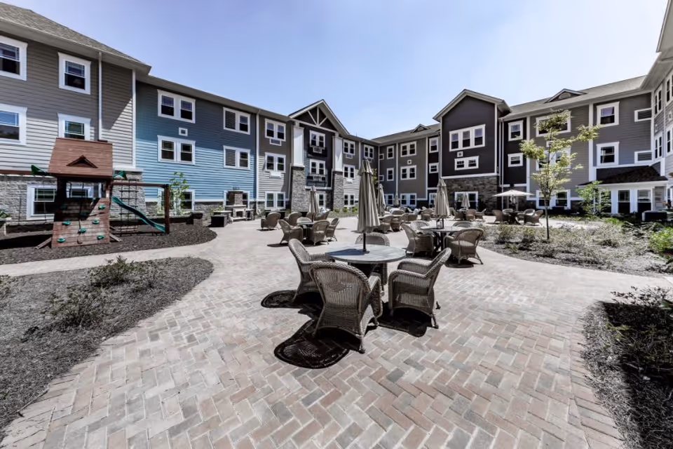 Outdoor courtyard area of The Phoenix at James Creek featuring multiple round tables with umbrellas and wicker chairs arranged on a brick-paved patio. Surrounding the patio are three-story residential buildings with multiple windows, and a small playground structure with a slide is visible on the left side. The sky is clear and blue.