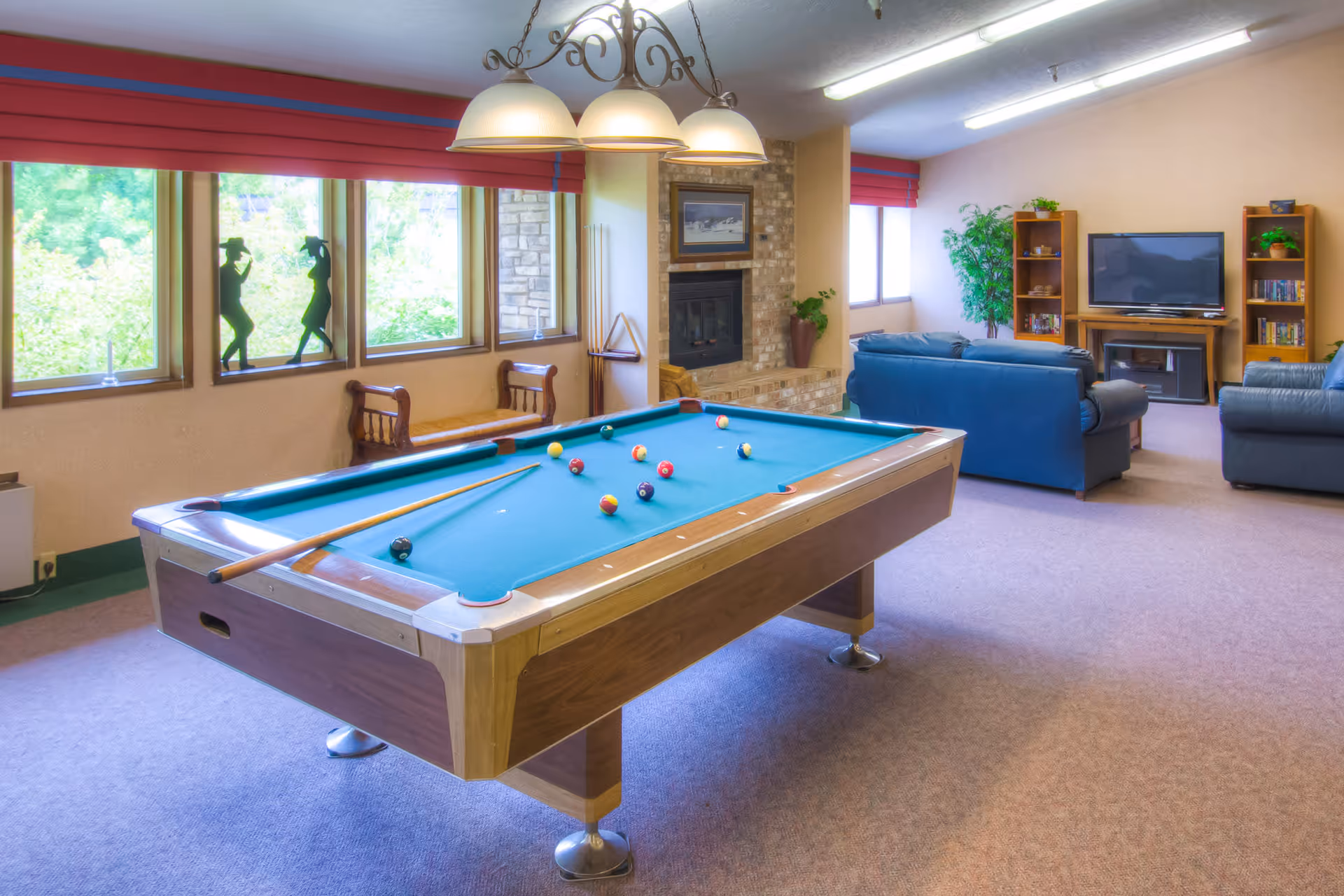 A recreation room with a pool table in the foreground, a cue stick resting on it, and billiard balls scattered on the table. In the background, there is a seating area with blue couches, a TV on a stand, bookshelves, a fireplace with a framed picture above it, and large windows with red blinds letting in natural light.