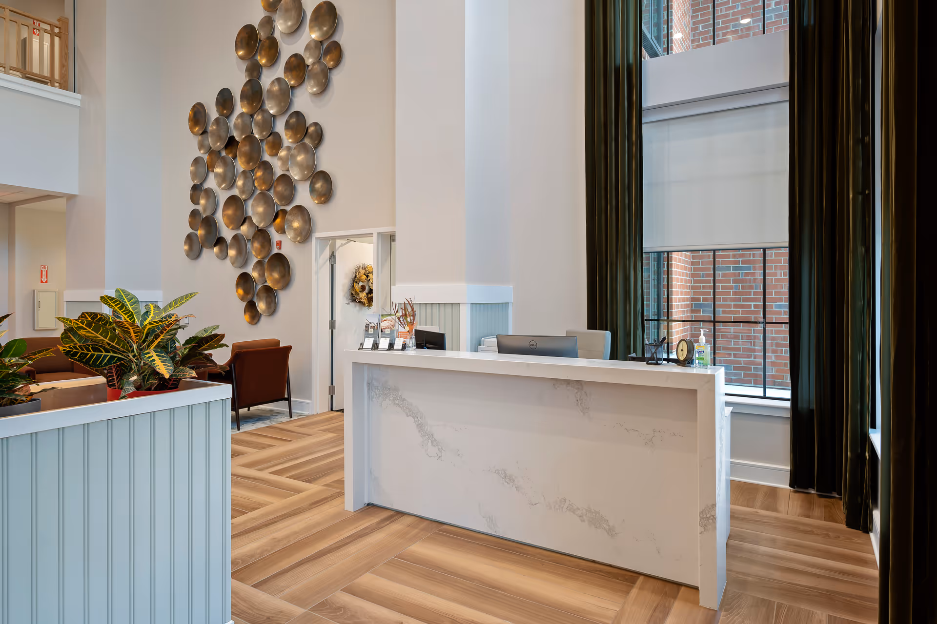 Reception area with a white marble front desk, two computer monitors, and a large window with dark green curtains. The floor is light wood with a herringbone pattern. On the left side, there are plants and brown chairs, and a wall decorated with a cluster of round metallic wall art pieces.