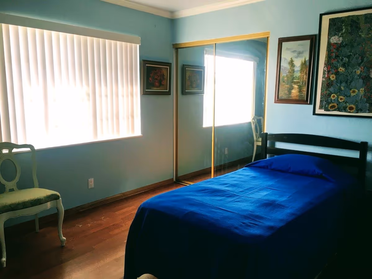 Simple bedroom with a single bed covered in a blue blanket, mirrored closet doors, a chair, and framed wall art.