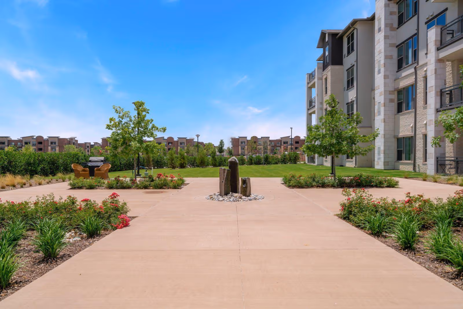 Sunlit landscaped courtyard with a central stone fountain, walkways, a seating area and an adjacent multi-story building under a blue sky.