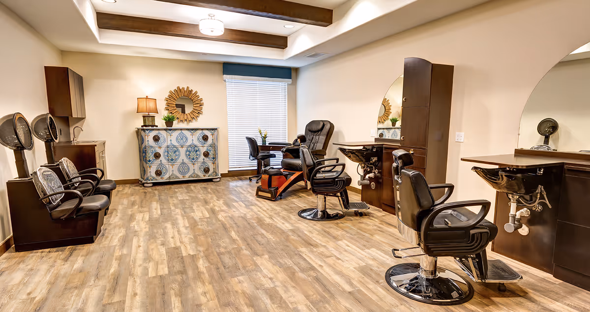 Interior view of a senior living facility's salon area featuring two black salon chairs with sinks, two hair drying chairs with hooded dryers, a pedicure chair, a small table with two chairs near a window with blinds, and a decorative chest with a lamp and plant on top. The room has wood flooring, beige walls, and exposed wooden ceiling beams.
