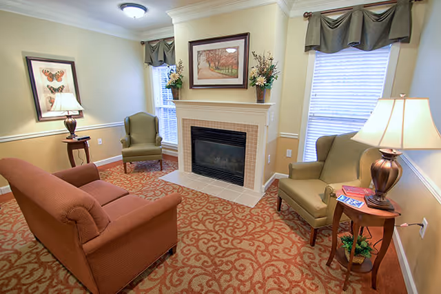 A cozy living room with a patterned carpet, a brown sofa, two green armchairs, a fireplace with a framed picture above it, two windows with valances, and two side tables each holding a lamp and decorative items.