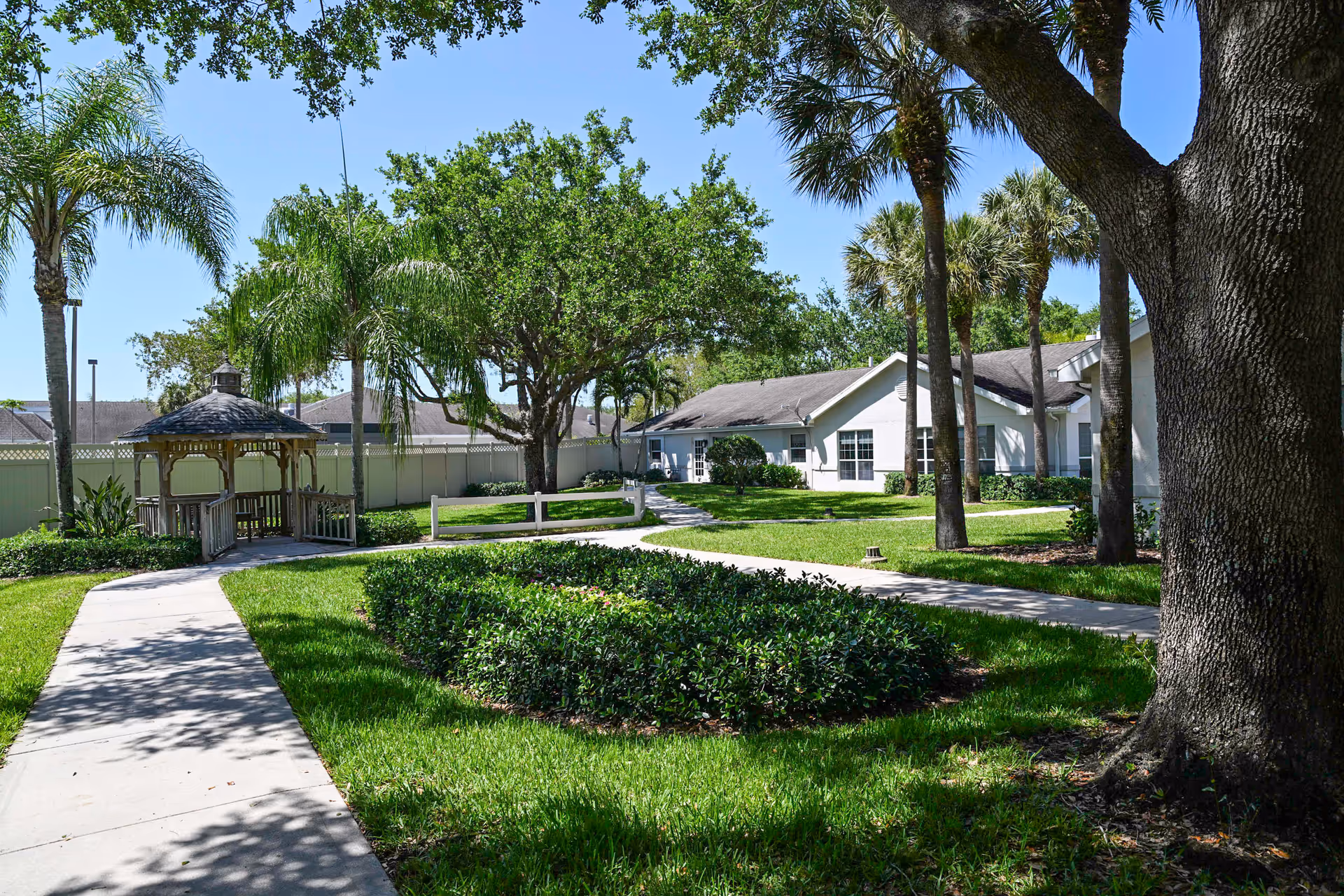 A sunny landscaped courtyard with a gazebo, paved walkways, palm trees, and single-story facility buildings.