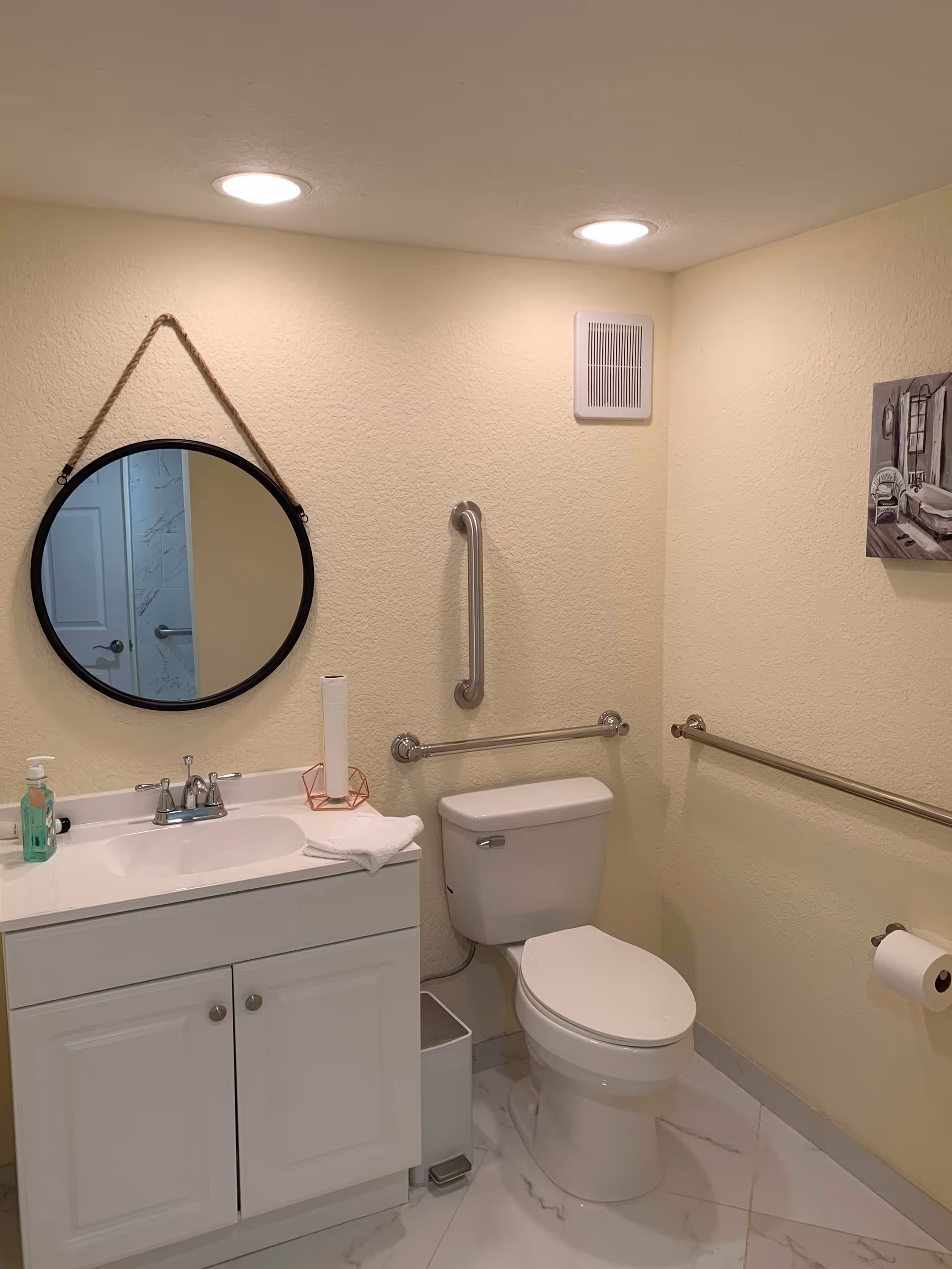 A clean and accessible bathroom with a white toilet, white vanity with sink, a round mirror hanging on the wall, grab bars next to the toilet, a paper towel roll on the vanity, a soap dispenser, and a small wall-mounted vent. The walls are light yellow and the floor has white tiles with gray veining.