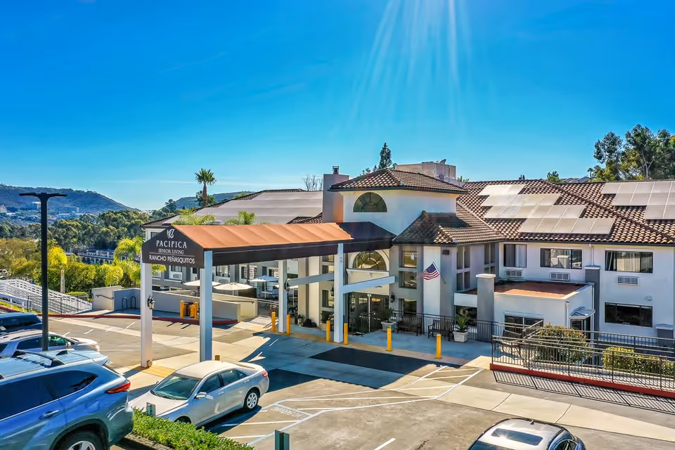 Exterior view of the Pacifica Senior Living Rancho Peñasquitos building with a covered entrance, parked cars in the parking lot, solar panels on the roof, and a clear blue sky with sunlight.