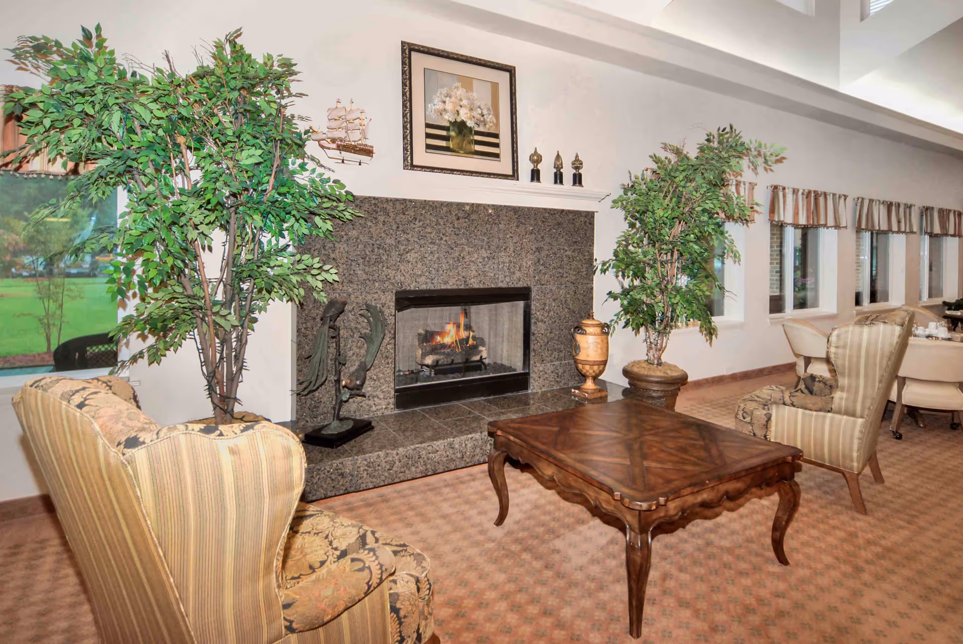 A cozy living room area with a lit fireplace surrounded by a granite mantel. Two patterned armchairs and a wooden coffee table are positioned in front of the fireplace. There are two large potted plants on either side of the fireplace, a decorative ship model, and framed artwork above the mantel. Windows with striped valances line the wall to the right, and a dining area with chairs and tables is visible in the background.
