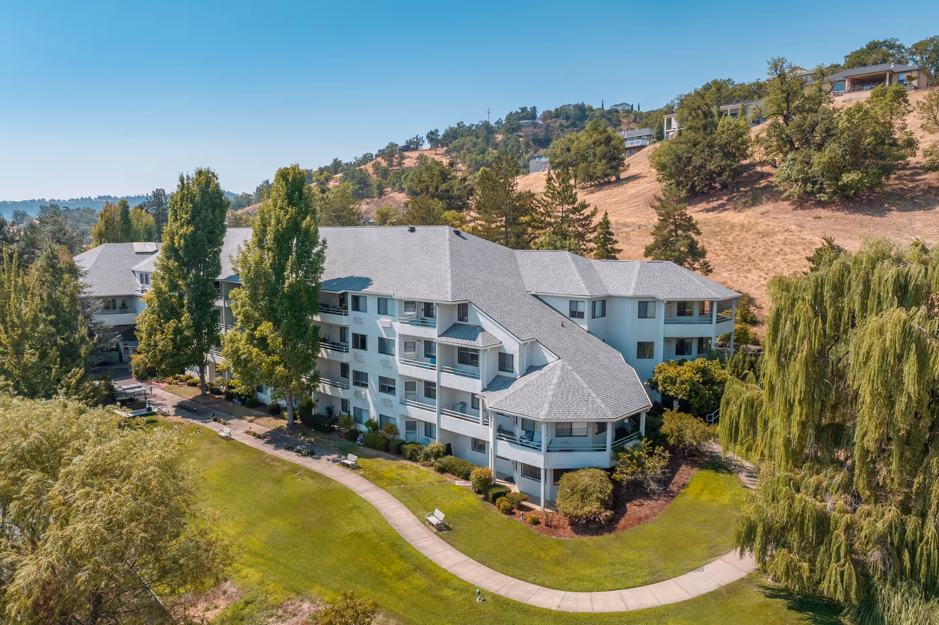 Aerial view of a multi-story senior living facility named Solista Roseburg surrounded by green lawns, trees, and a walking path with benches. The building is white with a gray roof and is set against a backdrop of hills with scattered trees and houses.