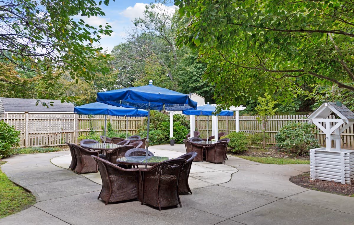 Outdoor patio area with several round glass tables surrounded by wicker chairs. Each table has a large blue umbrella providing shade. The patio is surrounded by greenery, trees, bushes, and a wooden fence. There is also a decorative white wooden wishing well on the right side.