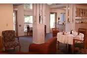Open communal dining and lounge area with round tables covered in tablecloths, upholstered chairs, and a kitchen pass-through.