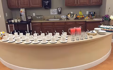 A curved buffet counter set with rows of teacups and saucers, glasses and pitchers of pink drink, plates of pastries and fruit bowls in front of cabinets and a sink.