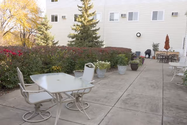 Outdoor patio area with several tables and chairs on a concrete surface, surrounded by potted plants and bushes. The background shows the exterior wall of a building with windows and some outdoor furniture including a grill and an umbrella.