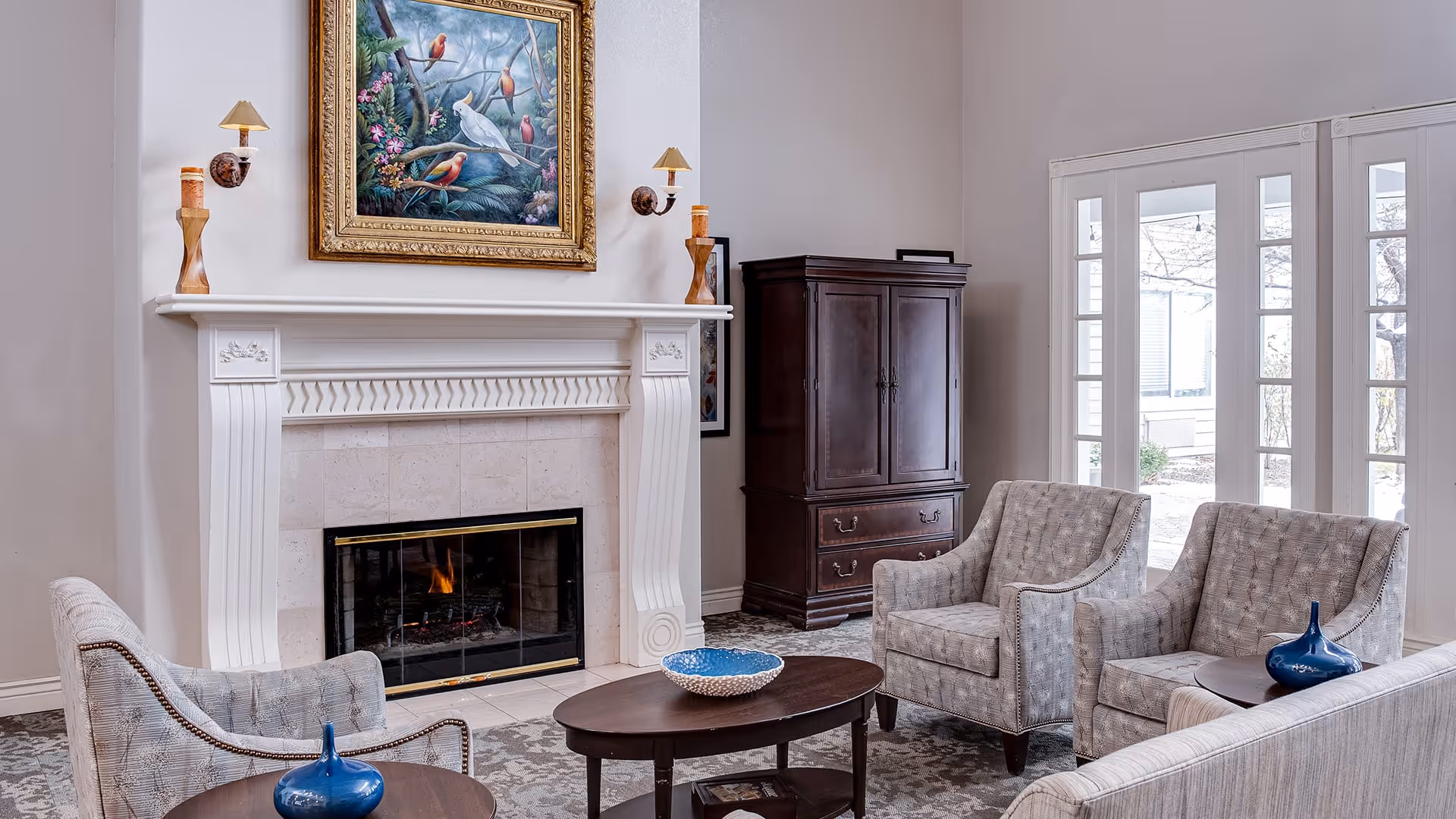 A cozy living room with a lit fireplace, two armchairs, a sofa, a dark wooden cabinet, and a coffee table with decorative items. Above the fireplace is a framed painting of colorful birds. Large windows with glass doors let in natural light.