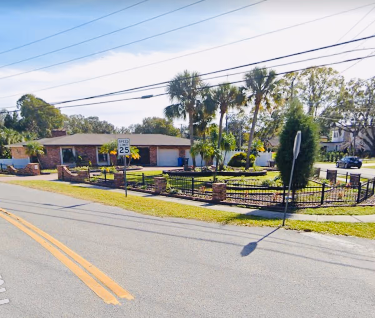 Street view of a single-story brick building with a fenced garden area featuring palm trees and other greenery. A speed limit sign of 25 mph is visible near the sidewalk, and power lines run overhead.