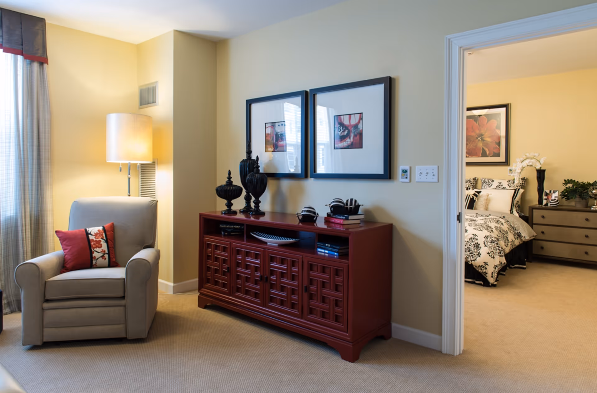 Cozy living area with an armchair, floor lamp, decorative red console and framed art, with a bedroom visible through an open doorway.